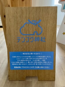 「テンゴク神社」の木製看板が写っています。看板には雲のイラストと「テンゴク神社」という文字が描かれており、その下には説明文が書かれています。説明文には、十国峠の安全と人々の幸せを願って「テンゴク神社」が誕生したこと、開雲絵馬に願いを込めることを勧める内容が記されています。絵馬は「TENGOKU CAFE」で購入できる旨も書かれています。看板は木目調で、周囲の雰囲気と調和しています。