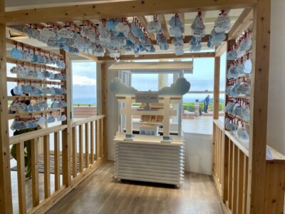 「TENGOKU CAFE」のすぐ隣にある「テンゴク神社」の内部が写っています。木製のフレームに多数の雲形の「開雲絵馬」が赤い紐で吊るされており、それぞれに願い事が書かれています。中央には小さな社が設置され、周囲の大きな窓からは明るい光が差し込んでいます。背景にはテラスと青い海が広がり、訪れた人々が景色を楽しんでいる様子が見えます。