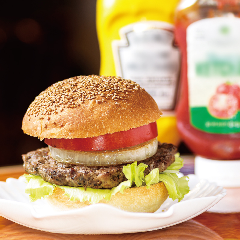 The Yokosuka Navy Burger at HONCH SHELL, with a thick patty, generously sliced onions, tomatoes, and lettuce nestled between sesame-seed buns, is served with bottles of mustard and ketchup.