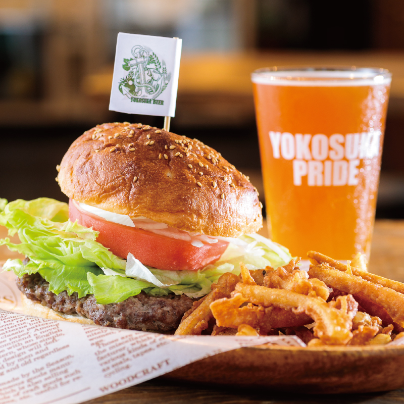 The Yokosuka Navy Burger, featuring a thick patty, lettuce, tomatoes, and onions tucked between buns, is accompanied by crispy French fries and a glass of craft beer poured at Yokosuka Beer