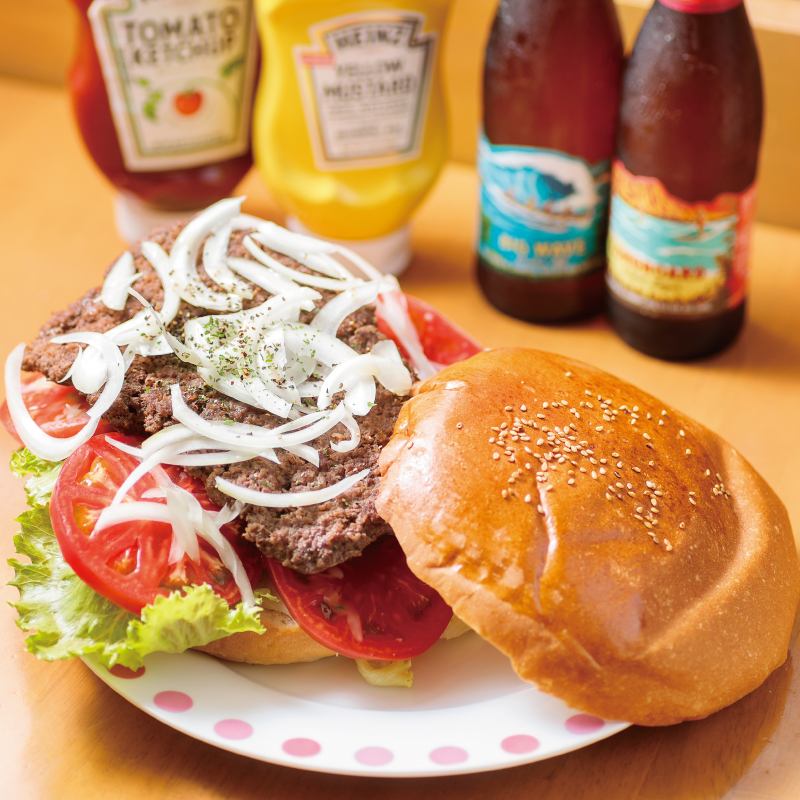 The Yokosuka Navy Burger at YOKOSUKA Shell features a large sesame-seed bun filled with lettuce, several slices of tomato, a large patty, and plenty of shredded onions, served with bottles of mustard and ketchup, and a well-chilled bottle of beer.