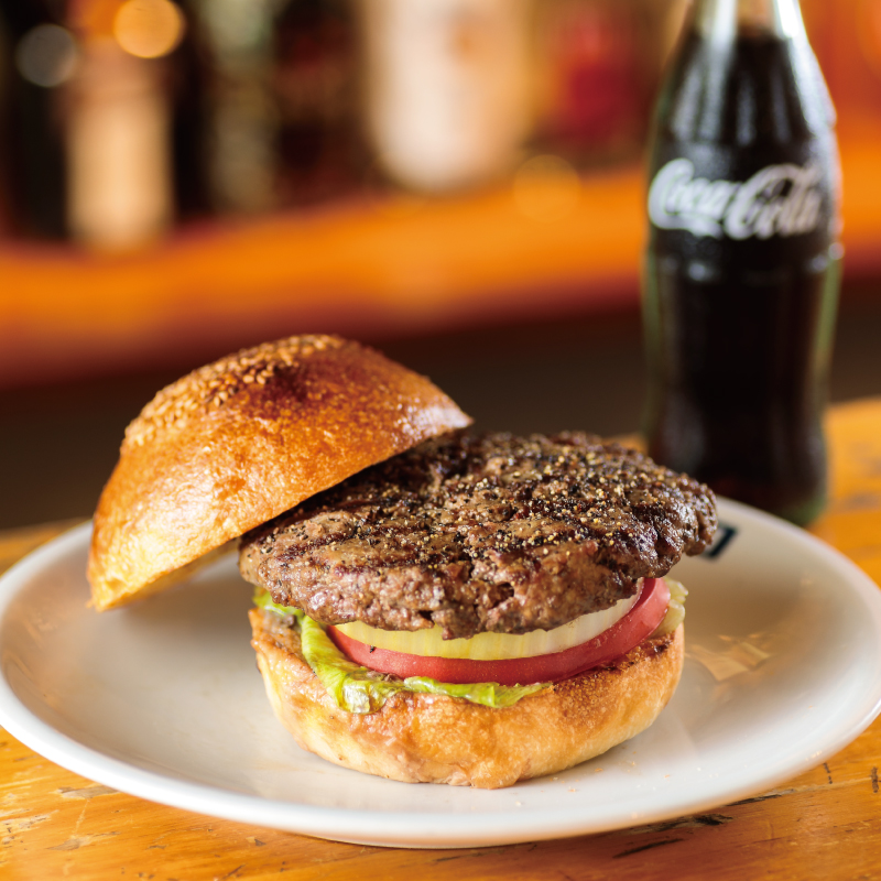 The tempting Yokosuka Navy Burger at TSUNAMI features a thick, toasted bun filled with lettuce, tomato, onion, and a large patty seared to perfection, served with a bottle of Coca-Cola.