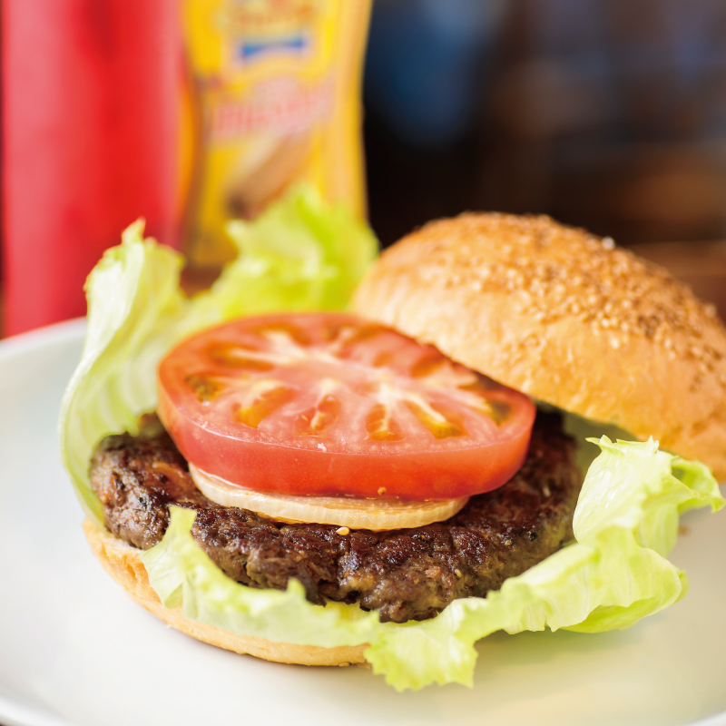 The Yokosuka Navy Burger at YOKOSUKA Shell, featuring a thick patty, onions, tomatoes, and lettuce tucked into sesame-seed buns, is served alongside bottles of mustard and ketchup.