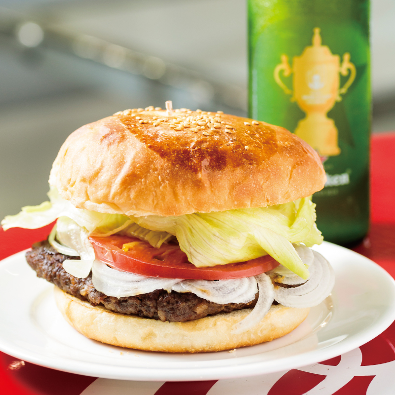 Yokosuka Navy Burger at Yokosuka Perry Gourmet Diner: A delicious hamburger with a sesame seed bun, perfectly grilled, topped with a juicy beef patty, onion slices, tomato, and lettuce. Served with a side of green beer bottle, creating a mouthwatering feast.