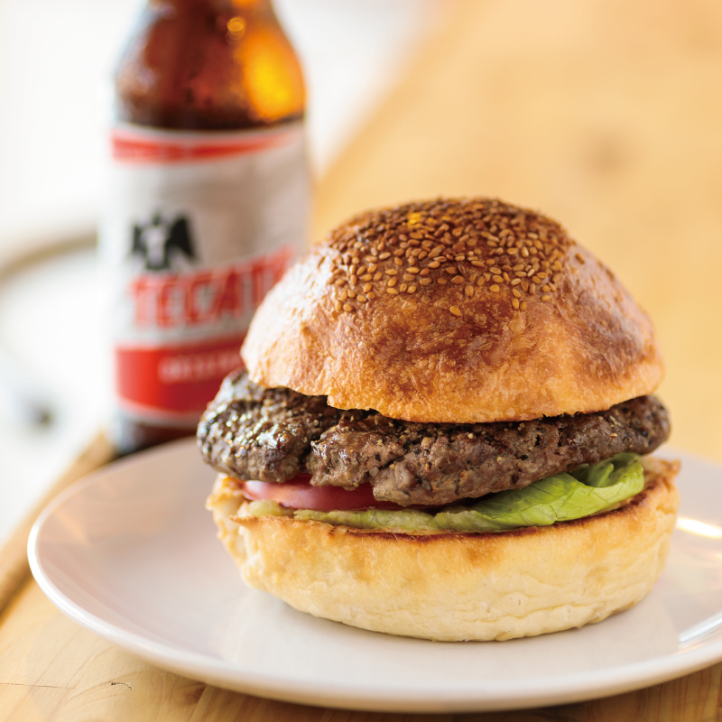 The enticing Yokosuka Navy Burger at SURF TACO features a thick, toasted sesame-seed bun filled with lettuce, tomatoes, and a large, thick patty with a perfect sear, served with a bottle of beer.