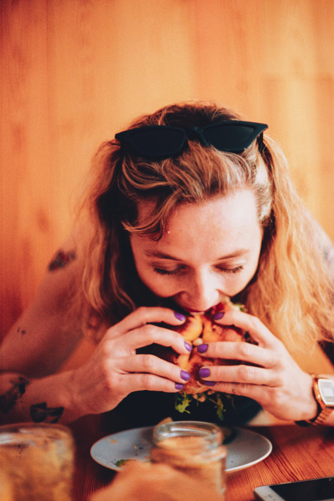  A blonde Caucasian woman enthusiastically grips a hamburger with both hands, taking a hearty bite.
