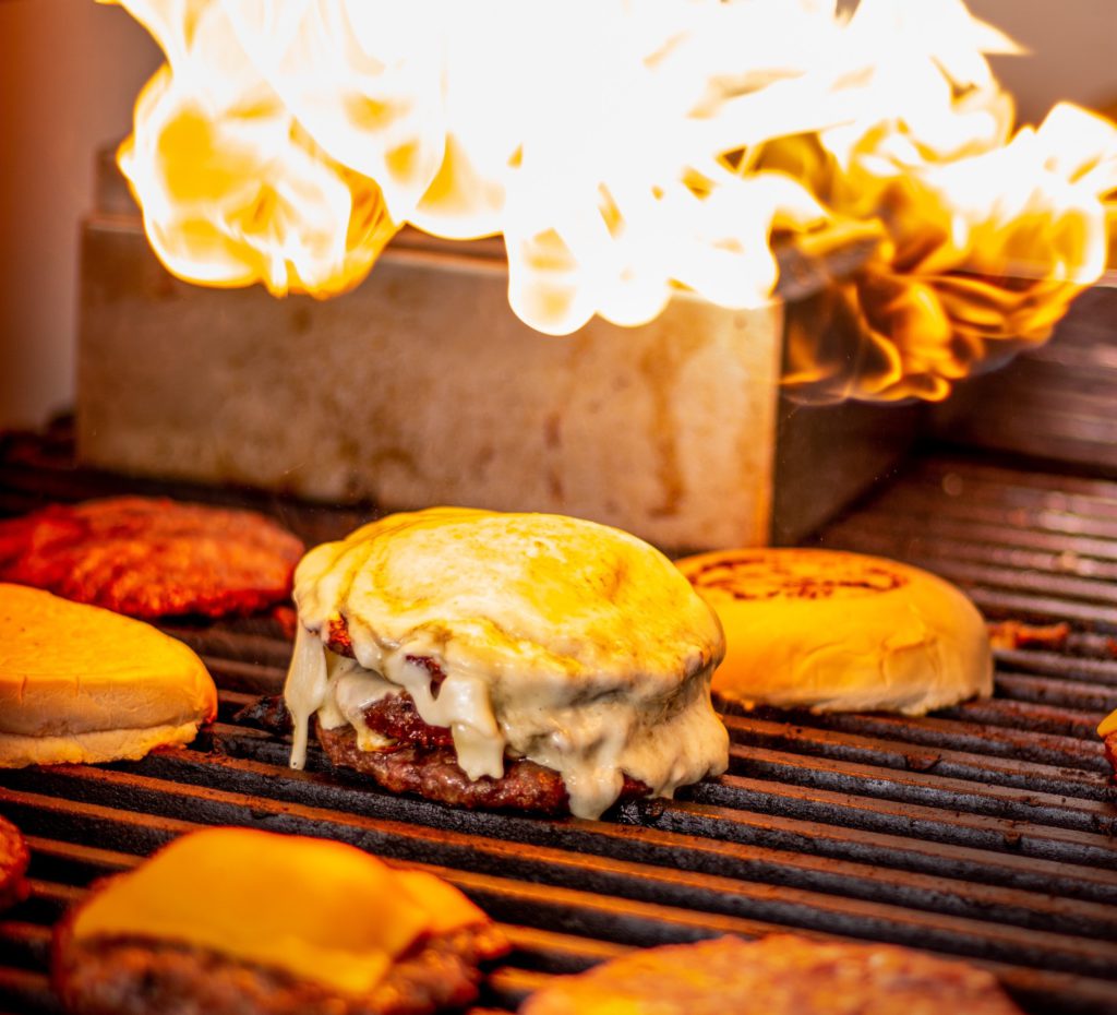 Three burger patties sizzle on a hot grill, crowned with melted cheese. Toasted buns line the sides as flames dance in the background.