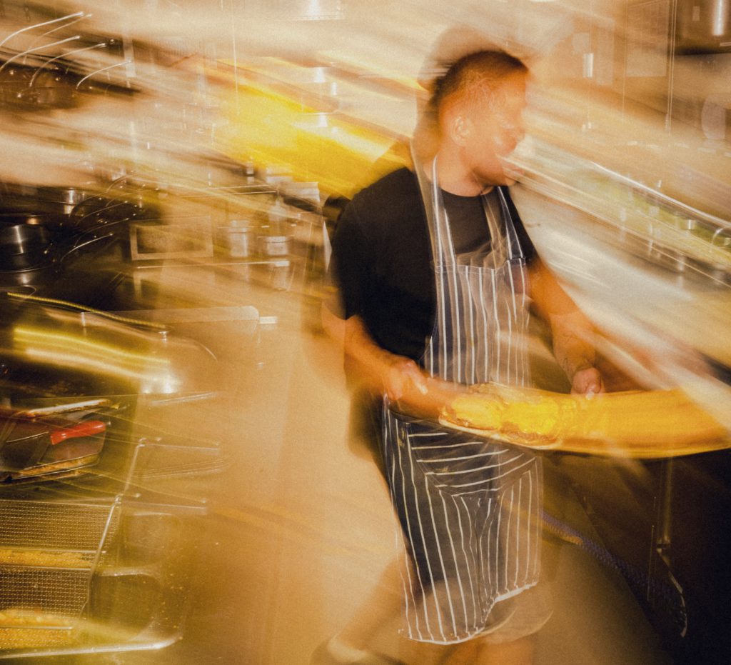 A chef, holding a plate with a burger, is bustling around the kitchen.