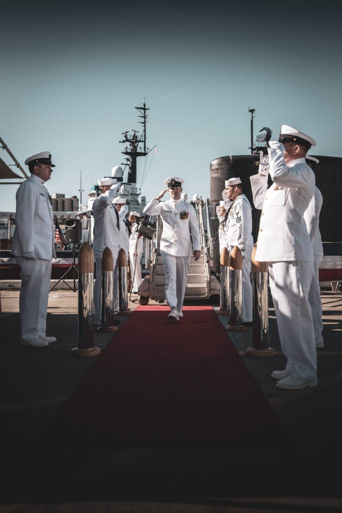 Navy soldiers stand in formation along the red carpet as a general, saluting, moves through the center. In the background, a battleship is visible.