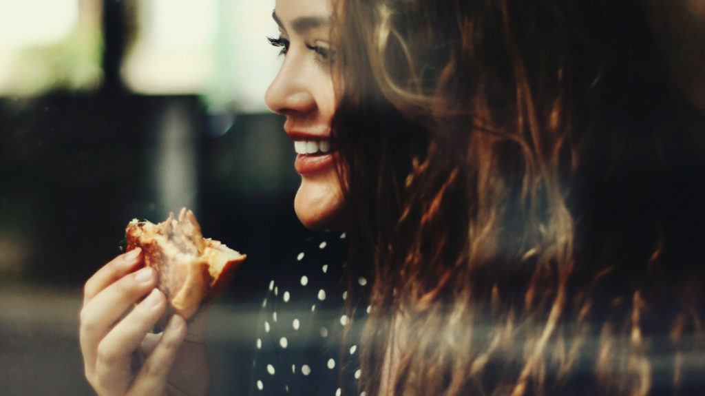 A brunette Caucasian woman, holding a partially eaten burger, is smiling.